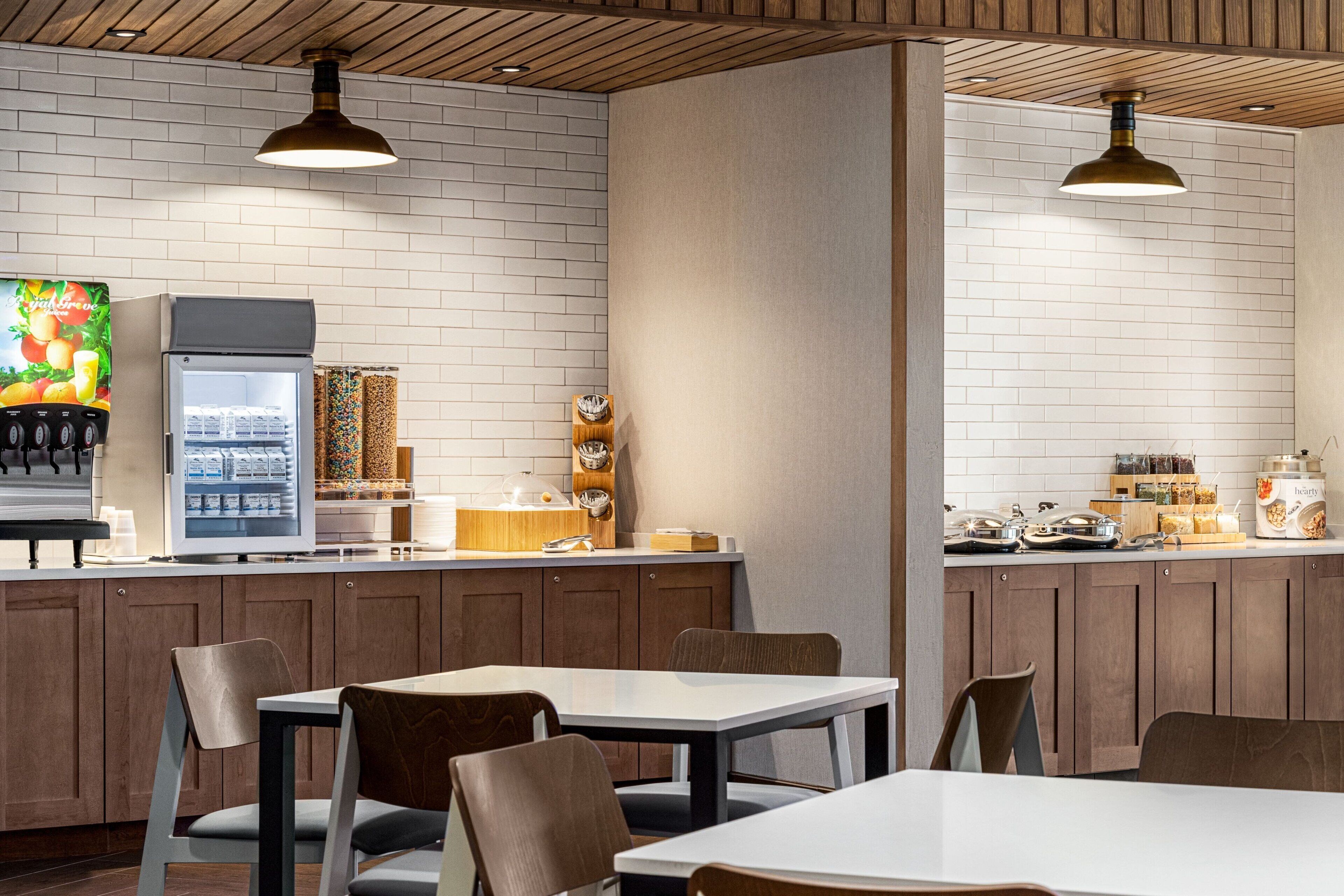 Restaurante moderno com parede de tijolos, teto de madeira e iluminação pendente. Há mesas, cadeiras, uma geladeira e uma máquina de café.