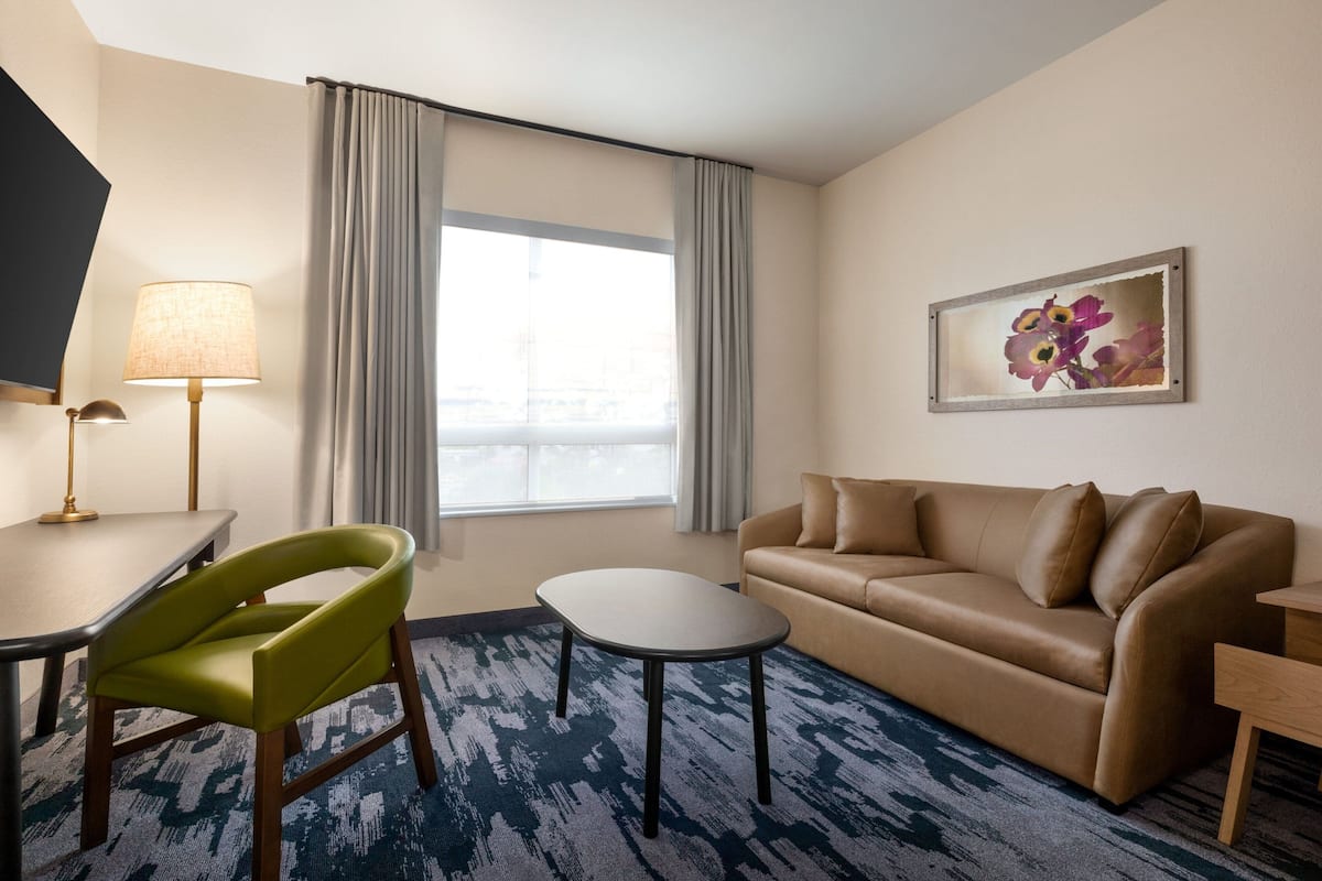Habitación de hotel con sofá, una mesita, lámpara, silla y ventana con cortinas.
