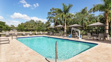 A rectangular swimming pool with a tiled deck, surrounded by lounge chairs and umbrellas, with palm trees and a fence in the background.