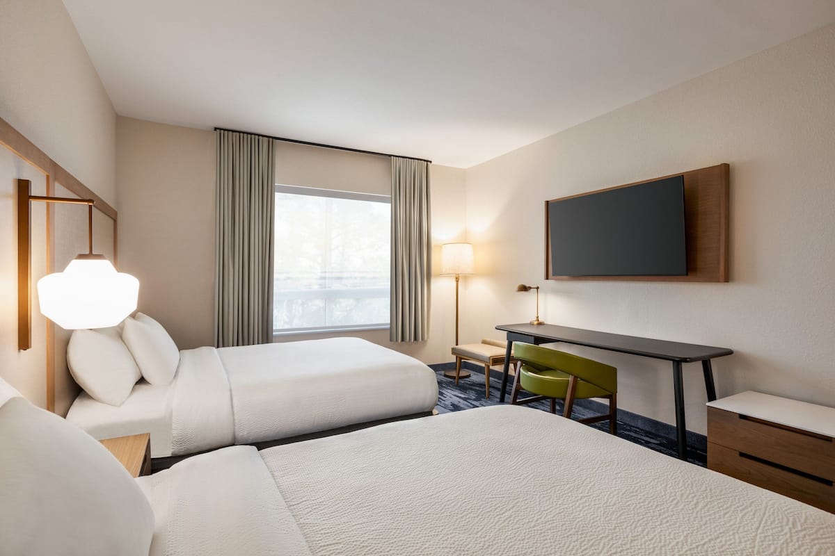 Habitación de hotel con dos camas, un escritorio, una silla, una lámpara y un televisor en la pared.
