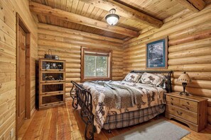 Um quarto de cabana de toras com uma cama, cômoda, prateleira com objetos decorativos, janela e um quadro na parede.