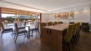 A spacious dining area with a long wooden table and multiple chairs, large windows offering a view of the outdoors, and a wall-mounted artwork. - TownePlace Suites San Luis Obispo (San Luis Obispo)