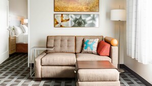 A modern living room with a sectional sofa, a small side table, and a floor lamp. There is a bed with a nightstand and a lamp in the background. - TownePlace Suites San Luis Obispo (San Luis Obispo)