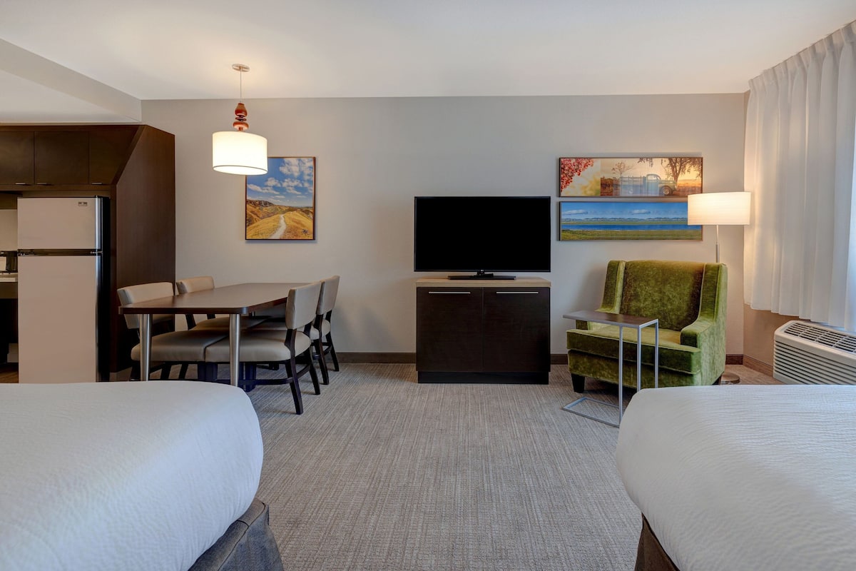 Une chambre d’hôtel avec deux lits, une table à manger avec des chaises, une télévision et des œuvres d’art accrochées aux murs.