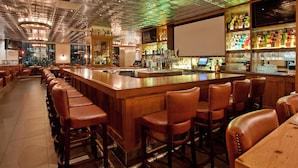 Una zona de bar con taburetes de madera, una barra larga y una variedad de botellas y vasos en estantes.