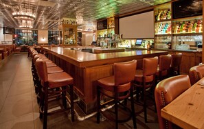 Uma área de bar com banquetas de madeira, um balcão comprido e variedade de garrafas e copos nas prateleiras.