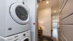 A laundry room with stacked washers and dryers, a wooden bench, a lamp, and a view of the hallway leading to a balcony. - Chic 2BR Condo w/ Open Living Space | Sleeps 4! (Nashville)