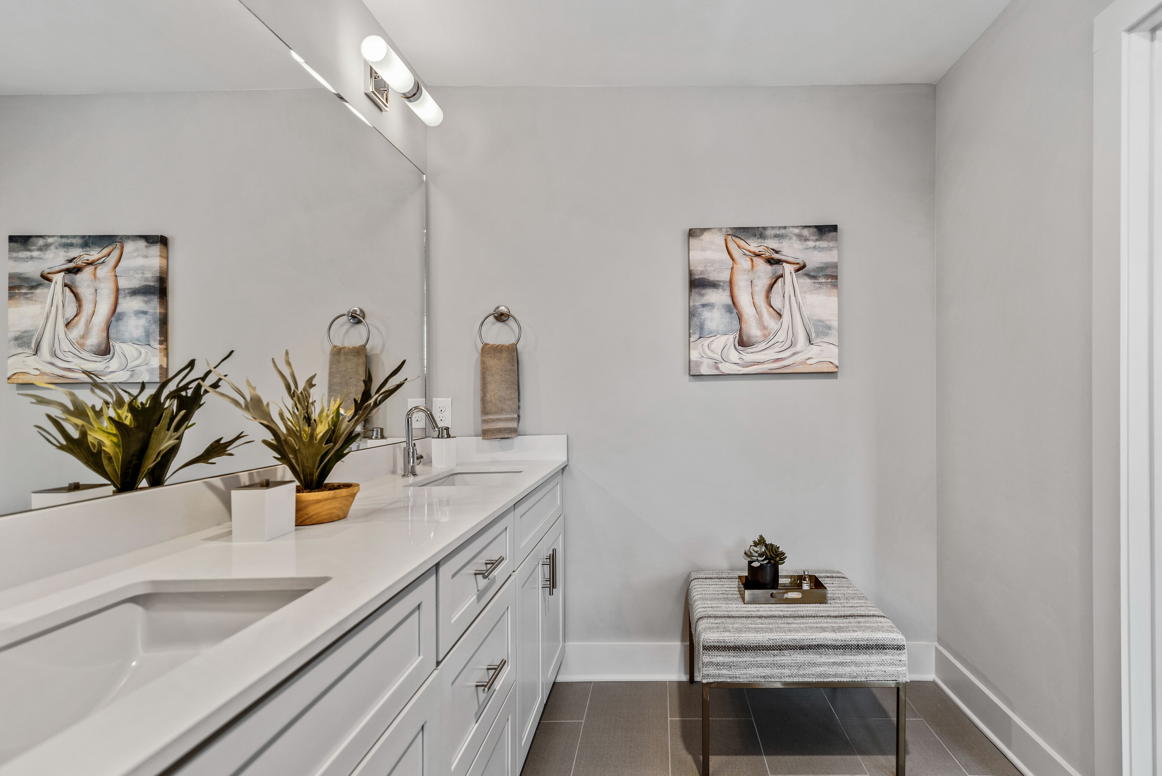 A modern bathroom with a large mirror, white cabinetry, a small bench, and two wall-mounted paintings.