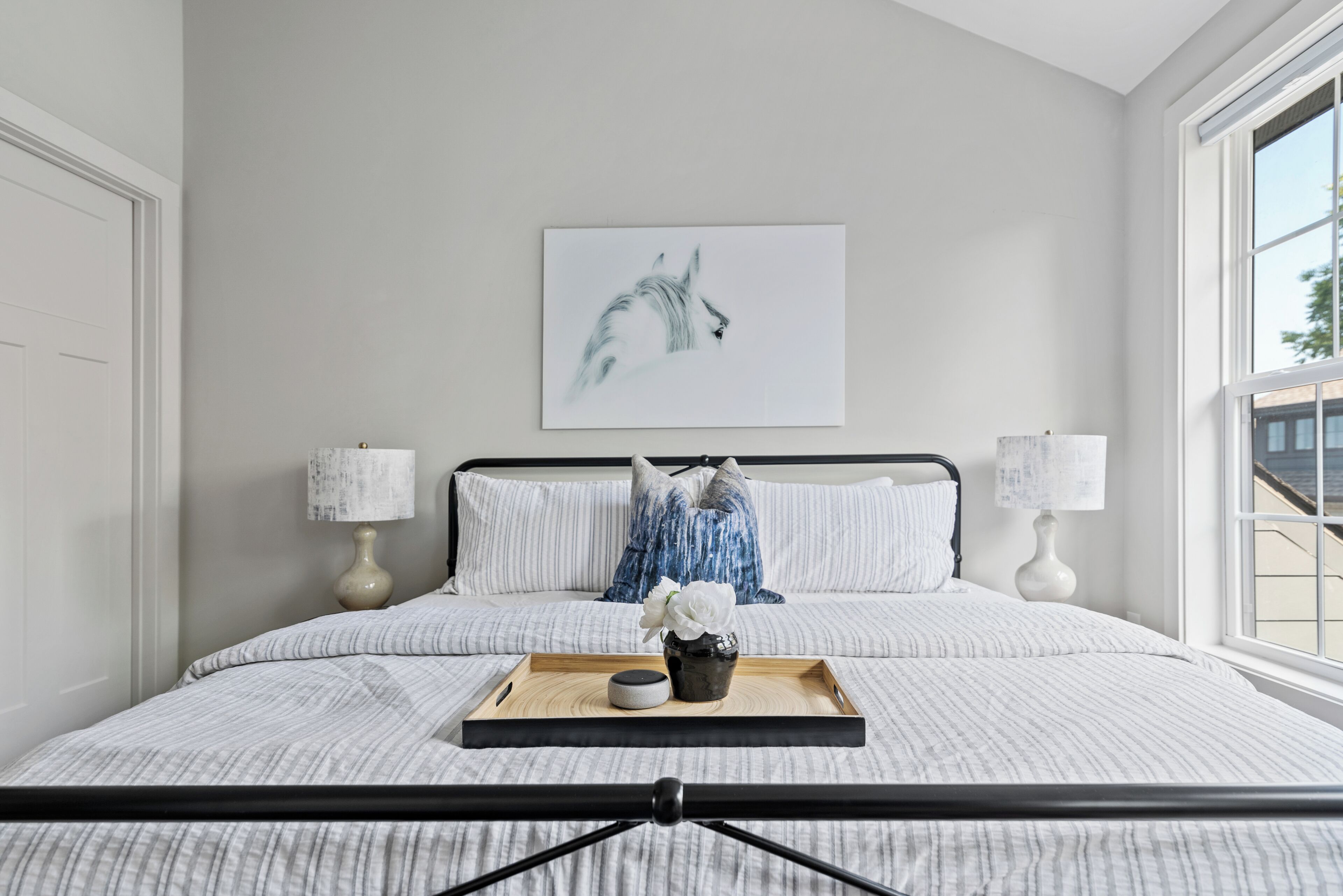 Ein Schlafzimmer mit Bett, Kissen, Nachttisch, Lampe, Tablett mit einer Vase und Blumen sowie einem Wandbild.