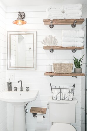 Un baño con lavamanos blanco, espejo y retrete. Se observan estantes de madera con toallas y una planta.