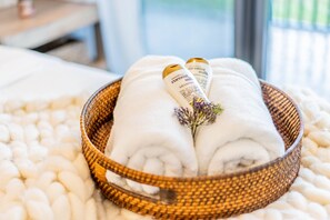 Una toalla blanca con una pequeña flor morada y un envase de acondicionador de leche de coco sobre una manta blanca con textura.