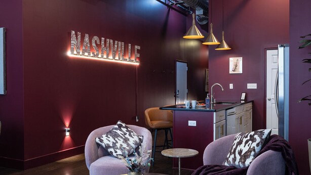 A modern living room with a red accent wall, a neon sign reading "Nashville," two armchairs with patterned cushions, a small round table, and pendant lighting. - Dazzling Downtown Loft in the Heart of Music City (Nashville)