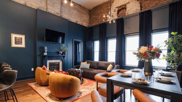 Un salón moderno con pared azul oscuro, techo de ladrillo, chimenea, sofá, mesa de comedor para cuatro personas y un ventanal con cortinas.