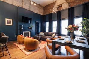 A modern living room with a dark blue wall, a brick ceiling, a fireplace, a sofa, a dining table set for four, and a large window with curtains.