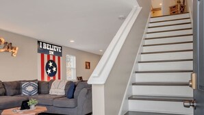 Ein modernes Wohnzimmer mit einem grauen Sofa, einem Couchtisch und einem gemusterten Teppich. Eine Treppe mit Holzwand verläuft in eine obere Ebene.