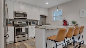 A modern kitchen with stainless steel appliances, white cabinetry, a large island with bar stools, and a window with blinds. - Honky Tonk Hideaway on Music Row (Nashville)