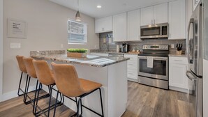 Una cocina moderna con armarios blancos, electrodomésticos de acero inoxidable, encimera de granito y una zona de bar con tres taburetes.