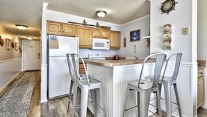 A kitchen with wooden cabinets, a white refrigerator, a microwave, and a bar area with metal stools. - Beachfront Oceanside Condo. Incredible view: Channel Marker  (Surfside Beach)