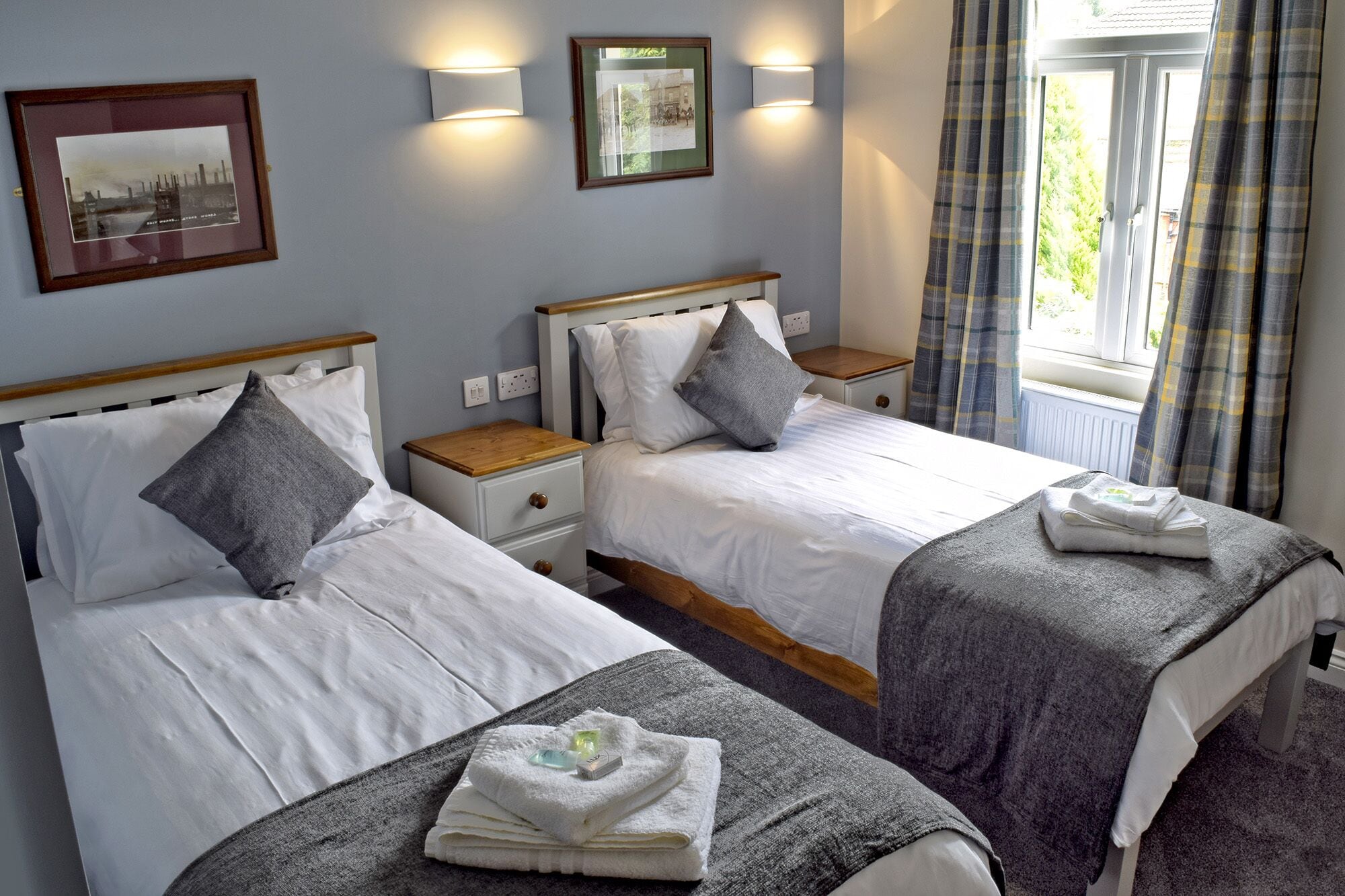 A hotel room with two beds, each with a wooden headboard, white bedding, and grey pillows. There are wall-mounted lights, a framed picture, and a window with curtains.