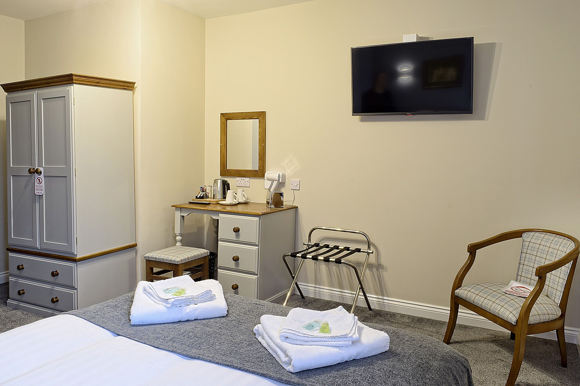 A hotel room with a bed, a desk with a mirror, a chair, a TV mounted on the wall, and a wardrobe.