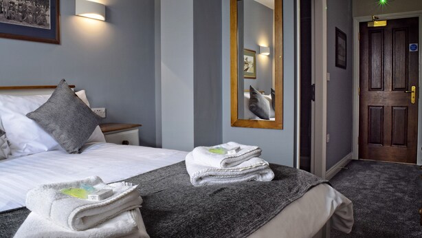 A hotel room with a neatly made bed, a stack of towels, a bedside table with a lamp, and a mirror on the wall. - Hanbury Turn (Bromsgrove)