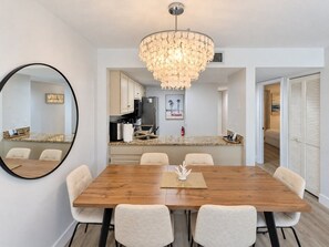 A modern dining room with a wooden table and chairs, a chandelier, and a kitchen in the background.
