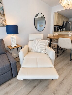 A modern living room with a recliner, a small table with a lamp, a dining area, and a kitchen in the background.
