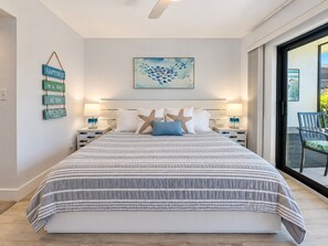 Une chambre avec un grand lit, deux tables de chevet, des coussins en forme d’étoiles de mer, une œuvre d’art accrochée au mur et une vue sur l’extérieur, visible à travers une porte vitrée.