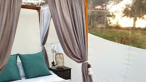 A tented bedroom with a bed, pillows, a bedside table with a lamp, a small plant, a woven basket, and a portable heater. - Africa's first Bubble Acomidation  (Piet Retief)