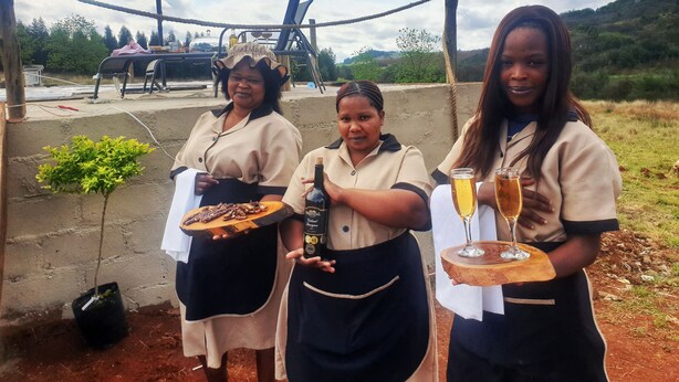 Three individuals outdoors, holding food and drinks, with a rope barrier and a small plant in the foreground. - Africa's first Bubble Acomidation  (Piet Retief)