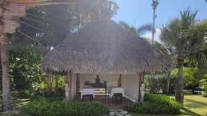 A thatched-roof structure with two massage tables and a statue, surrounded by lush greenery and a stone pathway.