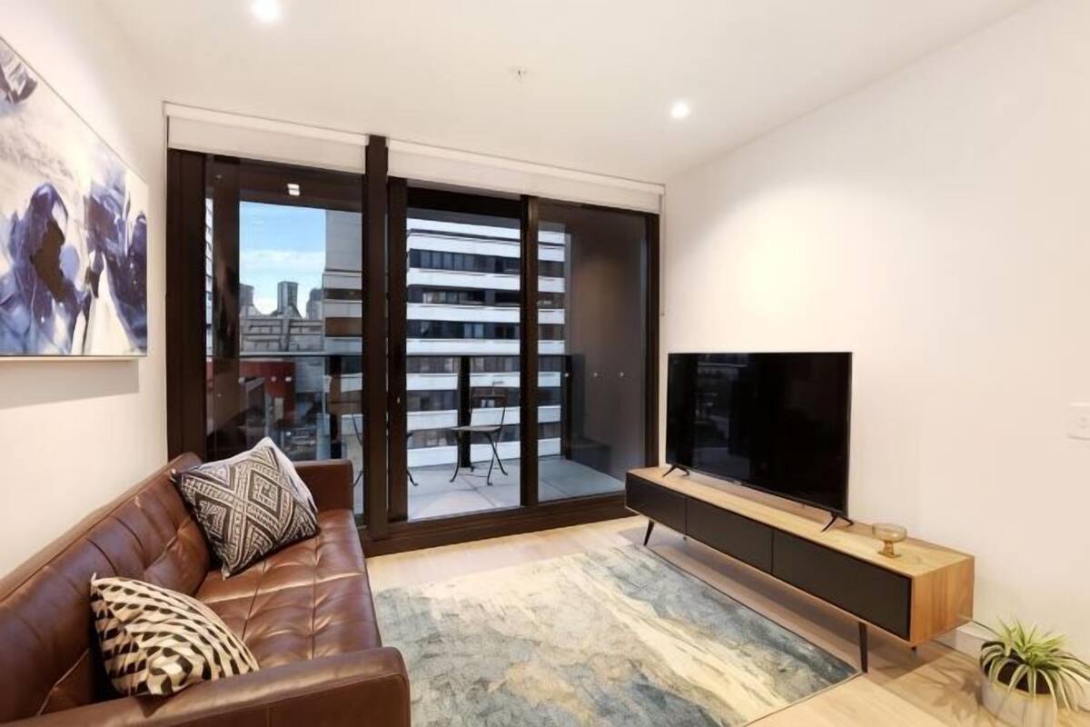 A modern living room with a brown leather sofa, a flat-screen TV, a wooden console table, and a large sliding glass door leading to an outdoor balcony.