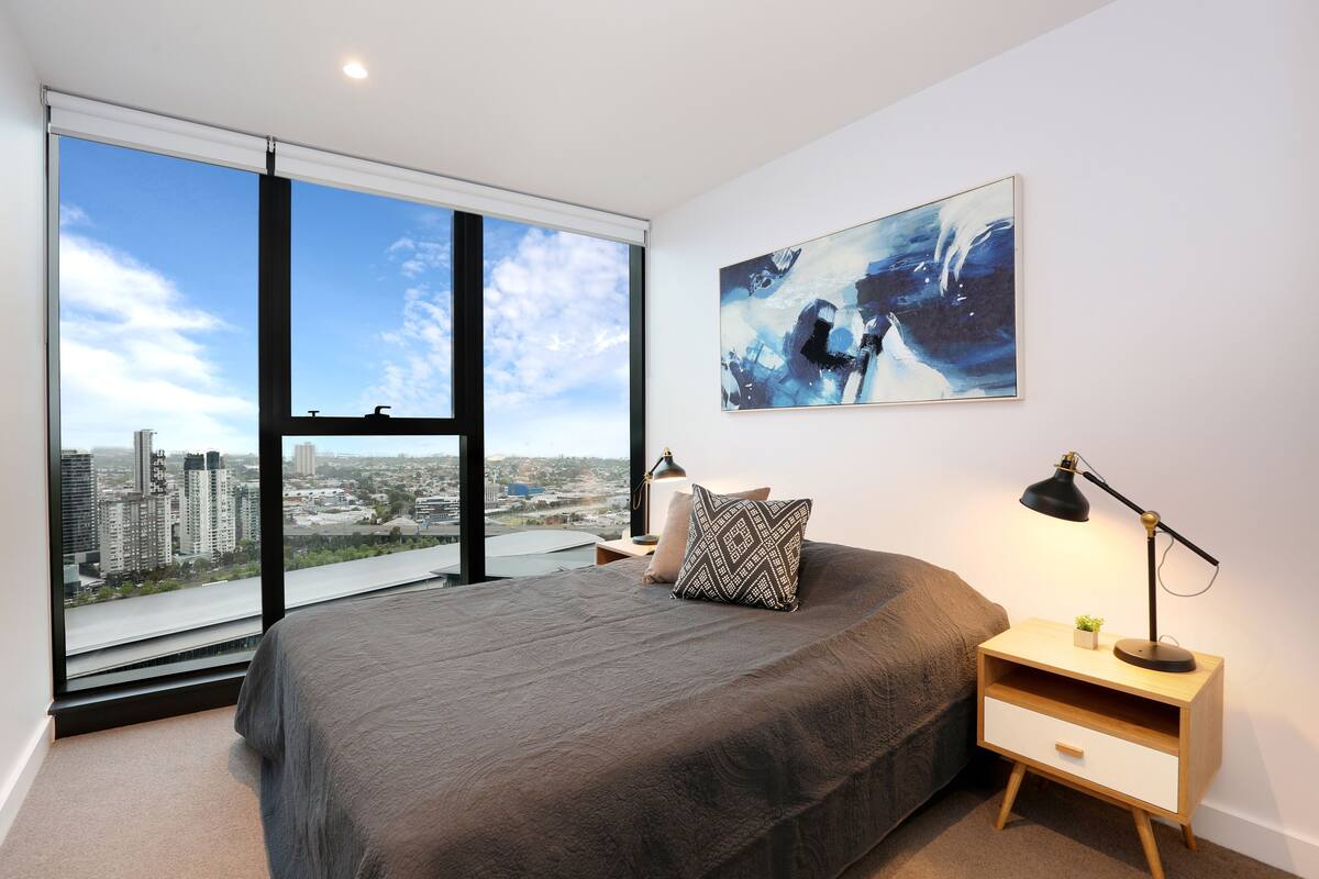 A bedroom with a large window, a bed, a nightstand with a lamp, and a painting on the wall.