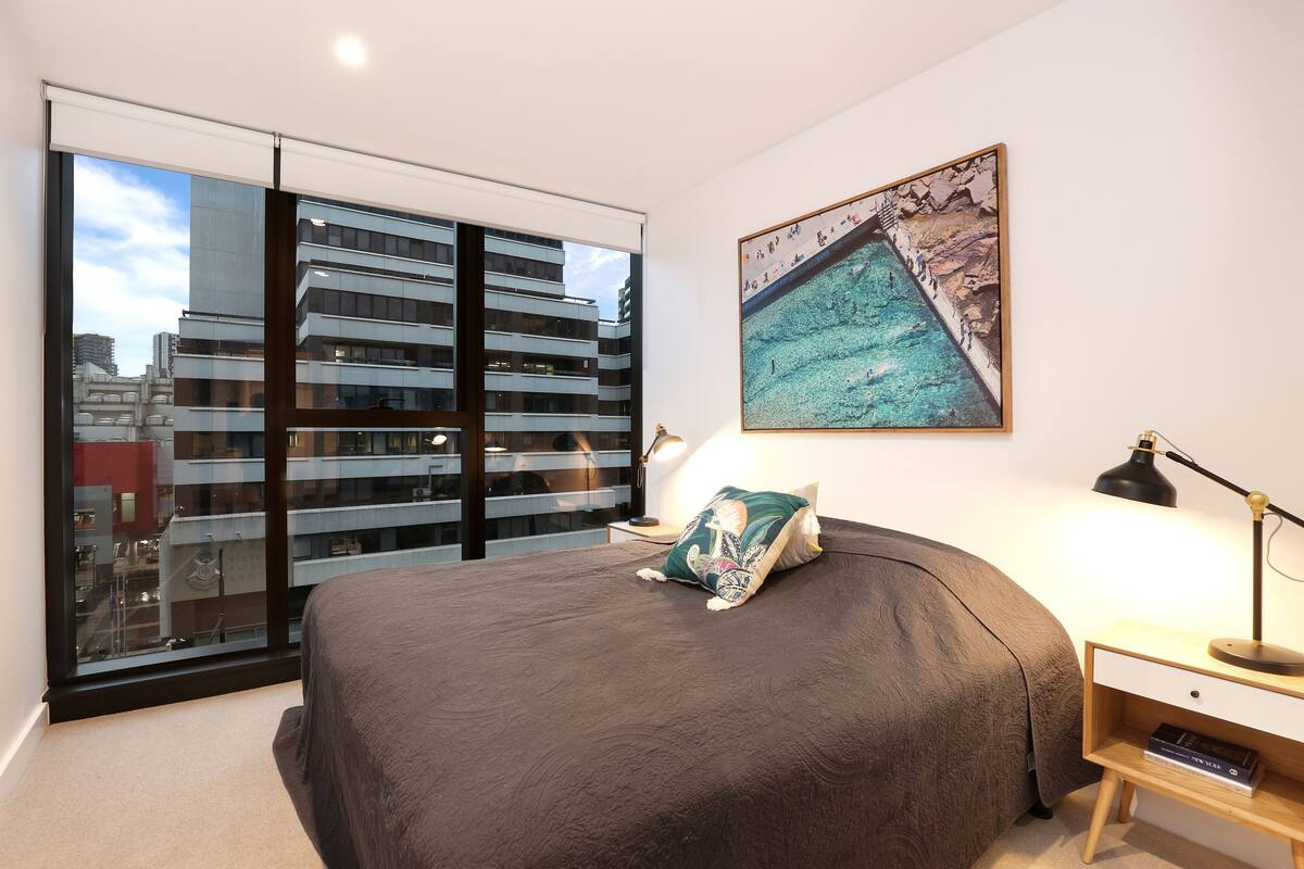 A bedroom with a large bed, a nightstand, a lamp, a framed picture, and a view of a cityscape through a sliding door.