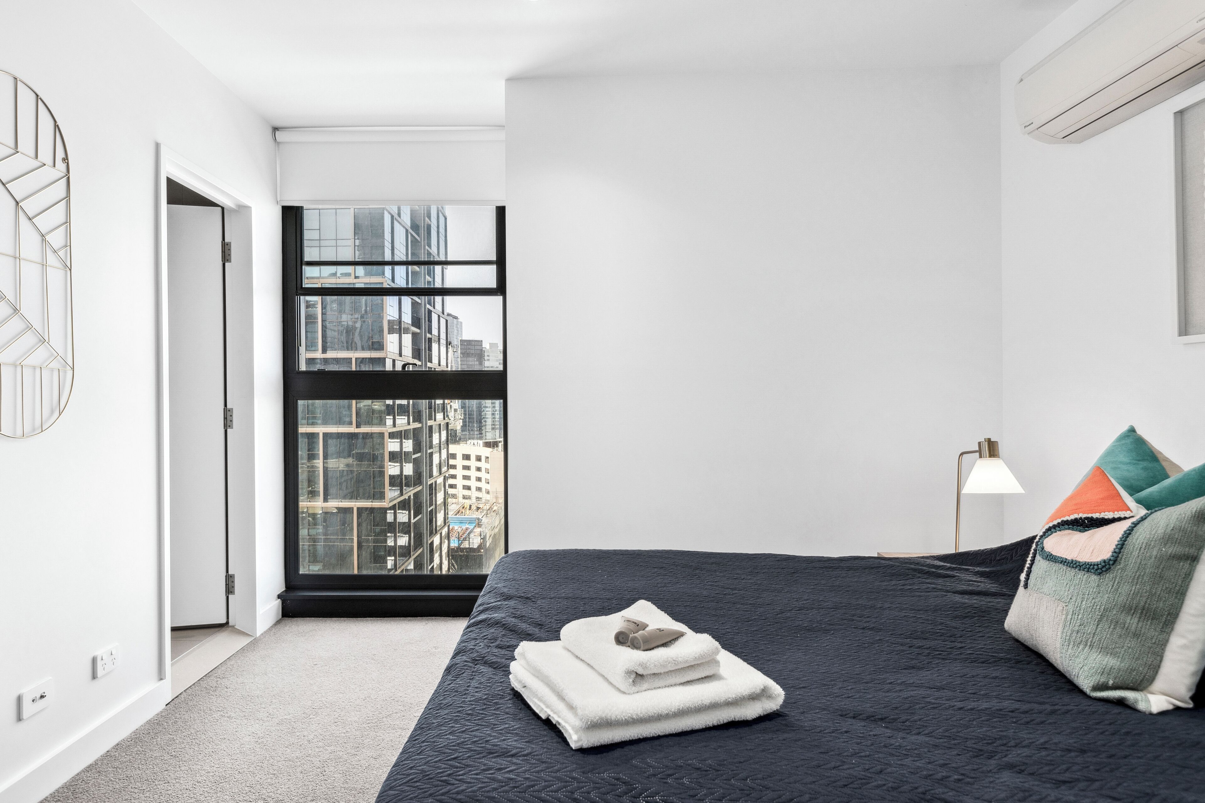 A modern bedroom with a large window, a bed with a dark quilt, and a view of a cityscape.