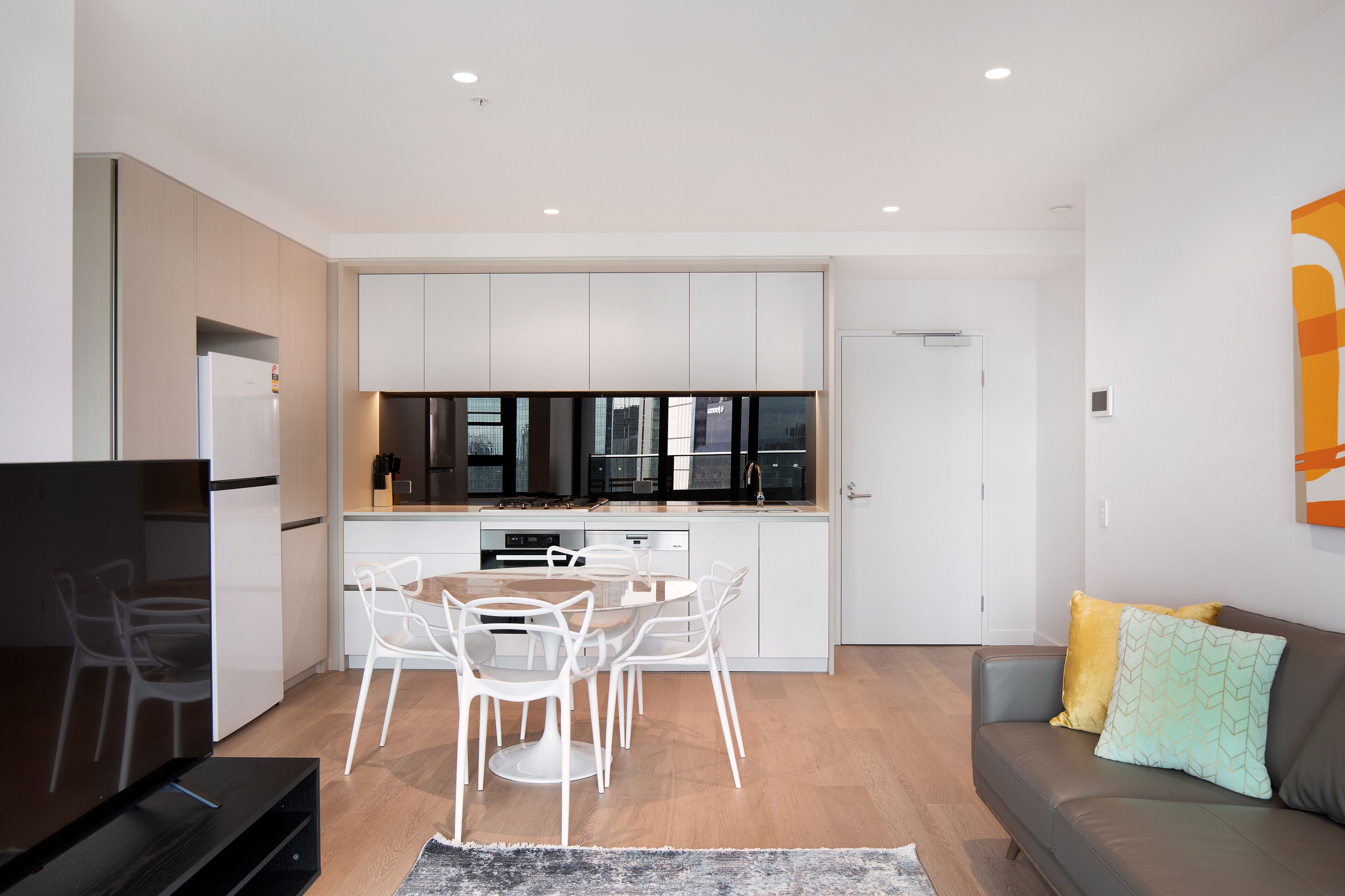 A modern kitchen with white cabinets, a dining area with white chairs, a gray sofa, and a TV.