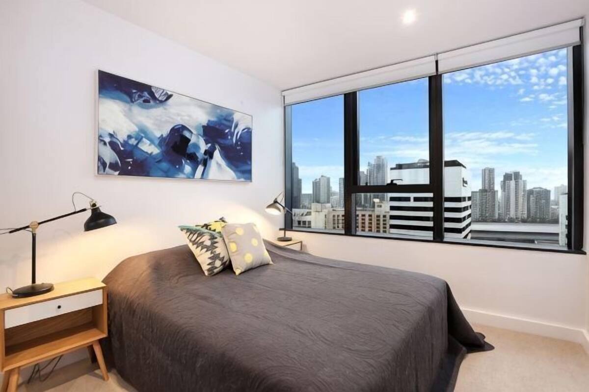 A bedroom with a bed, a nightstand, a lamp, a window with a city view, and a painting on the wall.