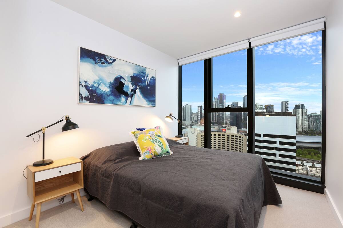 A bedroom with a large bed, a nightstand, a desk lamp, a colorful pillow, and a cityscape view through large windows.