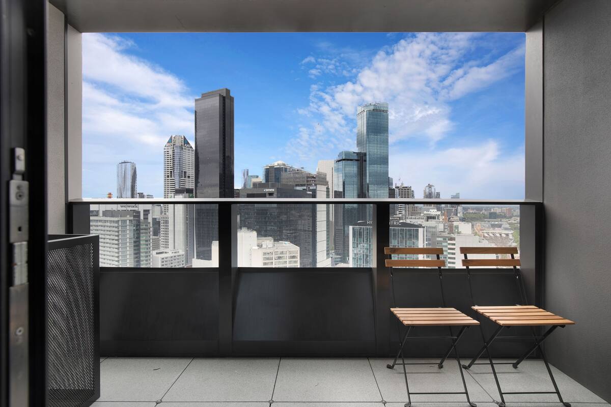 A balcony with a city view, two wooden chairs, and a metal trash bin.