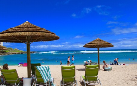 A beach with thatched umbrellas, green lounge chairs, and a view of the ocean.