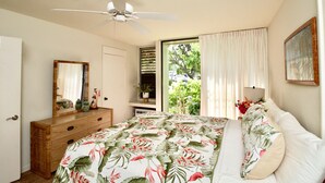 Une chambre à coucher comprenant un lit, un ventilateur de plafond, une commode, un miroir et une fenêtre avec des rideaux.