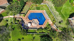 Vue aérienne d’une propriété dotée d’une piscine hexagonale, entourée d’un jardin et d’un espace patio aménagé avec du mobilier d’extérieur.