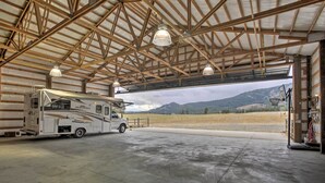 A spacious RV storage area with a large RV parked inside, a concrete floor, and a view of a mountainous landscape through an open garage door. - Rural Kettle Falls Bunkhouse w/ Airstrip & Trails! (Kettle Falls)