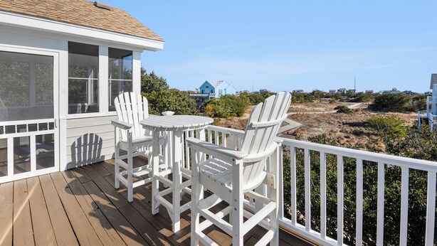 Une terrasse en bois, agrémentée de chaises Adirondack blanches et d’une petite table, offre une vue sur une plage à la végétation clairsemée et une maison bleue située au loin.