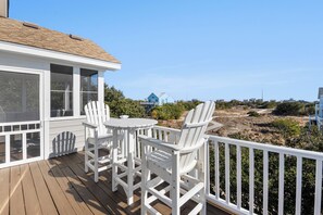 Une terrasse en bois, agrémentée de chaises Adirondack blanches et d’une petite table, offre une vue sur une plage à la végétation clairsemée et une maison bleue située au loin.