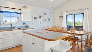 Une cuisine moderne avec des meubles blancs, une table à manger en bois et une vue sur l’extérieur grâce à de grandes fenêtres.