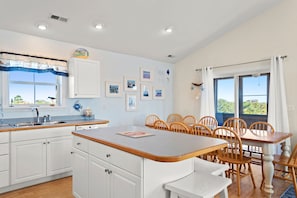 A modern kitchen with white cabinets, a wooden dining table, and a view of the outdoors through large windows.