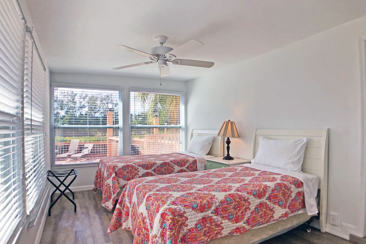 Une chambre avec deux lits, un ventilateur de plafond, une fenêtre avec des stores et une vue sur l’extérieur.