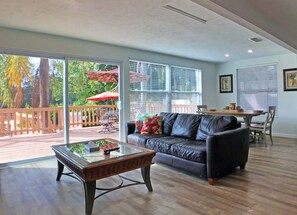 A spacious living room with a leather sofa, a glass coffee table, and a wooden dining table. Large windows provide ample natural light and a view of the outdoors.