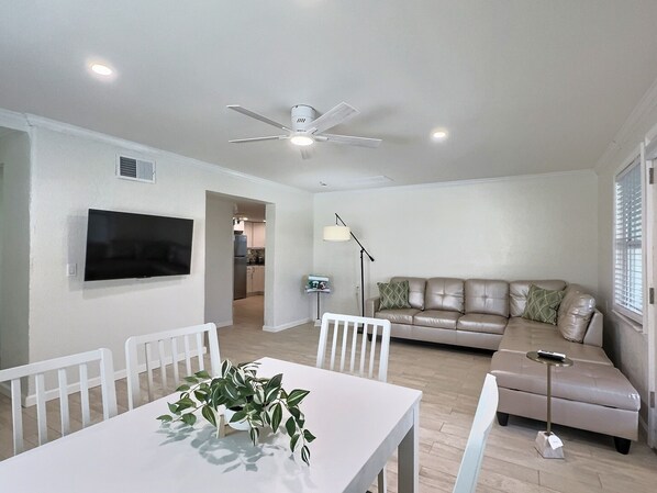 A modern living room with a white ceiling fan, a flat-screen TV mounted on the wall, a beige sofa, and a dining table with chairs.